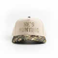 A baseball cap with a cream-colored crown and 'HE'S HUNTING' embroidered on the front, paired with a camouflage-patterned visor. A 'REALTREE' brand patch is visible on the brim.