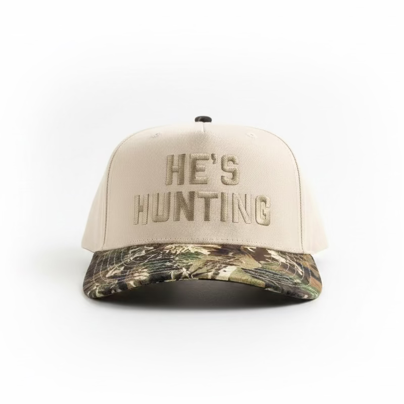 A baseball cap with a cream-colored crown and 'HE'S HUNTING' embroidered on the front, paired with a camouflage-patterned visor. A 'REALTREE' brand patch is visible on the brim.