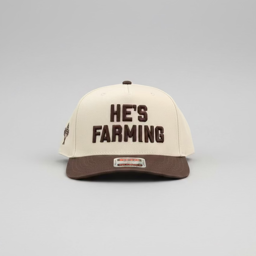 A baseball cap resting on a bed of dry grass and hay. The cap has an off-white crown with dark brown text reading 'HE'S FARMING' on the front, a dark brown visor, and a small dark brown logo on the side.