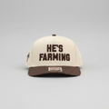 A baseball cap resting on a bed of dry grass and hay. The cap has an off-white crown with dark brown text reading 'HE'S FARMING' on the front, a dark brown visor, and a small dark brown logo on the side.